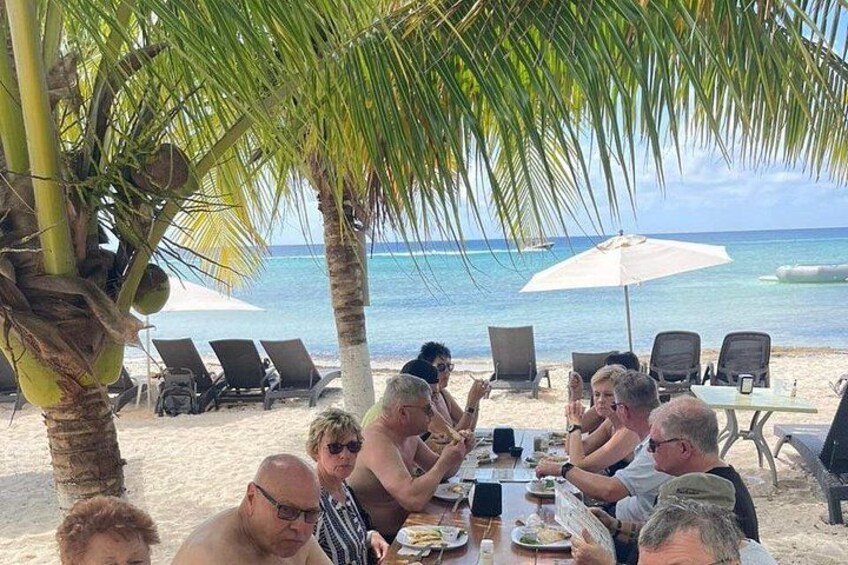 A group of people dining at a beachside table with palm trees and umbrellas in the background. 2
