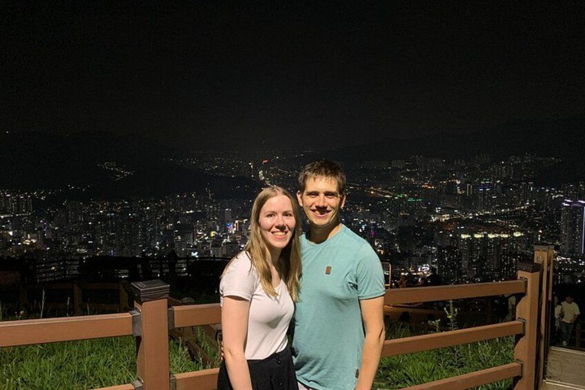 Two people standing on a balcony with a cityscape at night in the background. 3