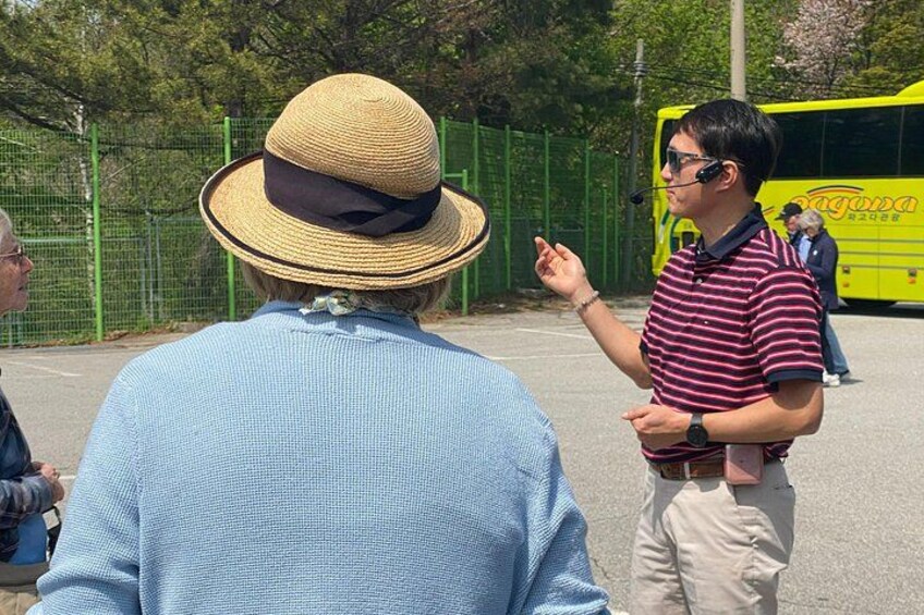 A person in a straw hat and striped shirt gesturing, a yellow bus, and a green fence in the background. 5