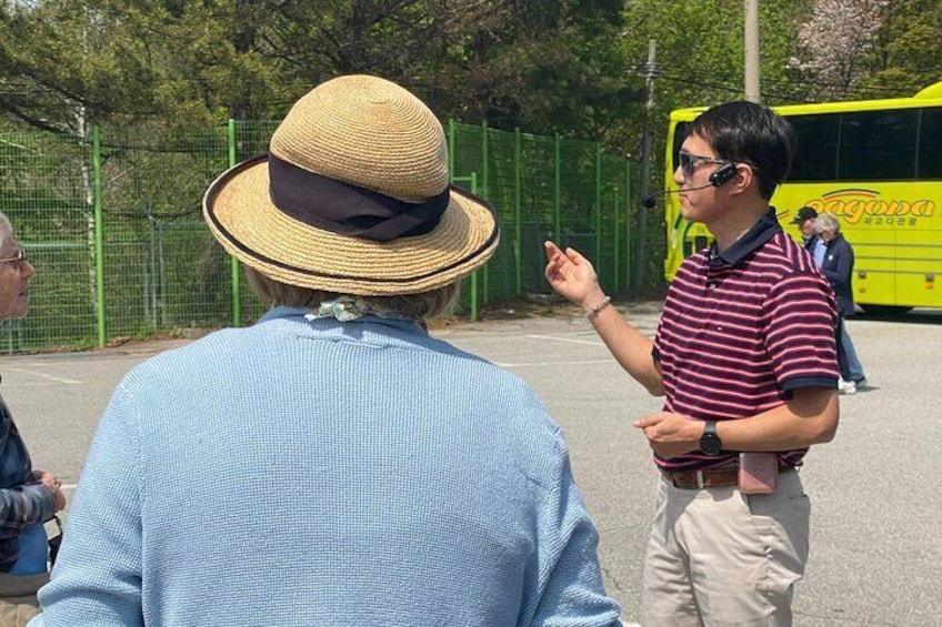 A person in a straw hat and striped shirt gesturing, a yellow bus, and a green fence in the background. 5