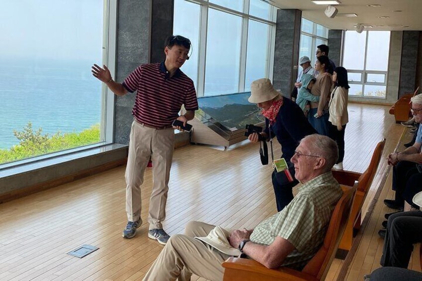 A man standing and gesturing in a room with large windows overlooking a body of water. 4