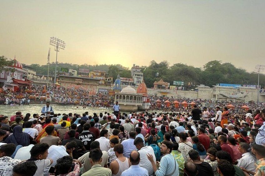 A crowded riverside with people gathered, a temple, and a bridge in the background. 5