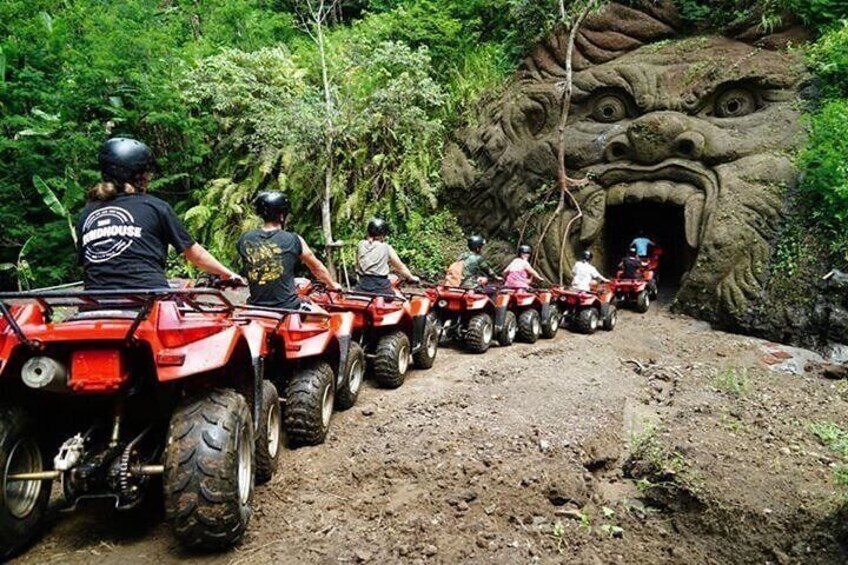 A group of people riding ATVs on a dirt path through a forested area with a large, carved rock face. 1