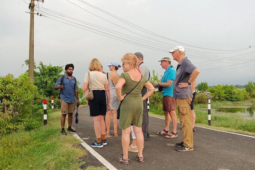 A group of people standing on a paved path beside a body of water, with greenery and a utility pole in the background. 5