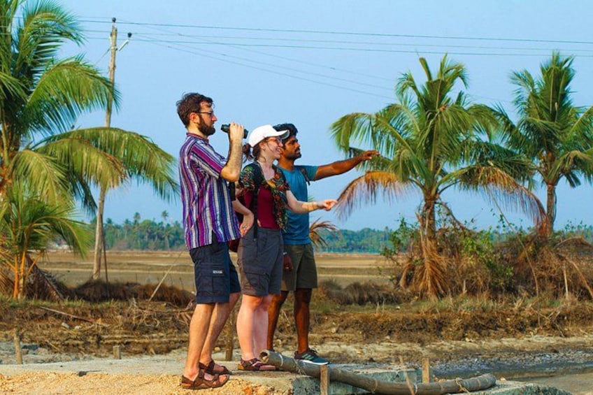 Three people standing outdoors, one holding a microphone, palm trees, and a rural landscape. 1