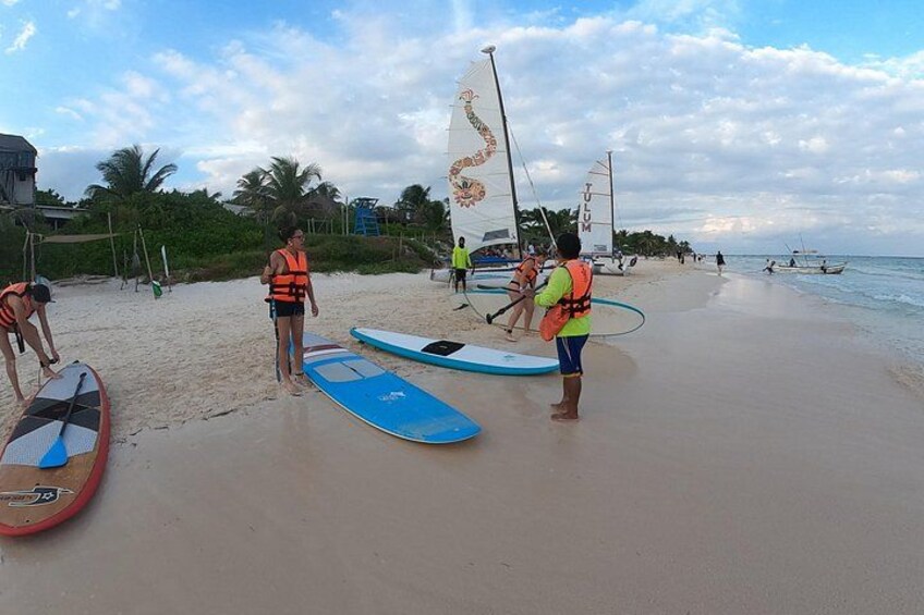 Una playa con gente preparándose para actividades acuáticas, incluyendo un velero y tablas de surf. 2