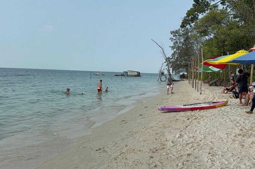 A beach with people swimming, a boat in the distance, and a colorful umbrella on the sand. 5