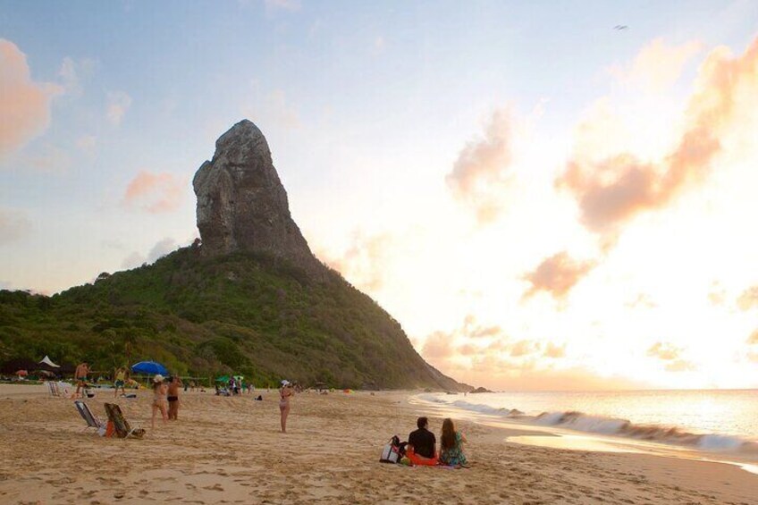 A beach with people sunbathing, a prominent rock formation, and the ocean. 1