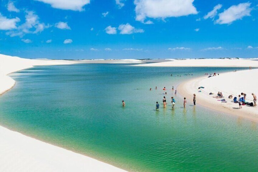 A beach with clear turquoise water, people swimming and sunbathing, and white sand dunes in the background. 1