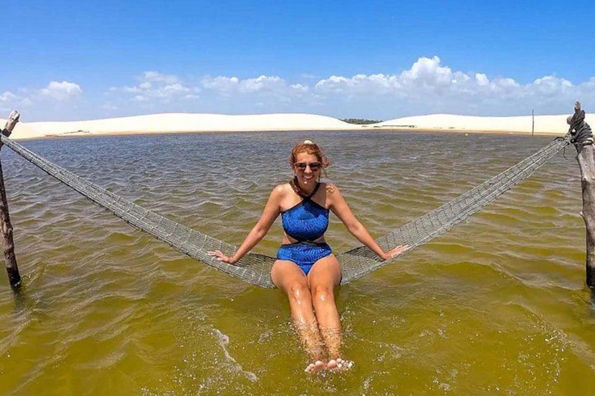 A person in a blue swimsuit sitting on a hammock in a body of water with sand dunes in the background. 2