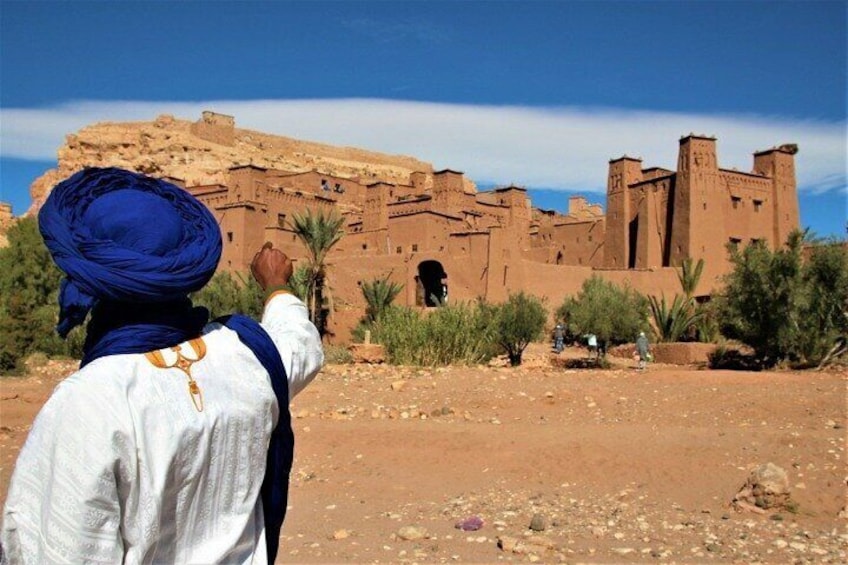 A person in traditional attire pointing at a large, ancient mud-brick building with multiple towers and arches. 1