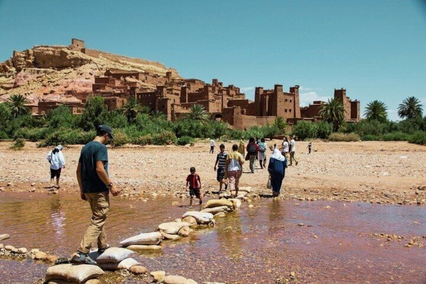 A group of people crossing a shallow water body with stepping stones, a large earthen structure in the background, and a clear sky. 5