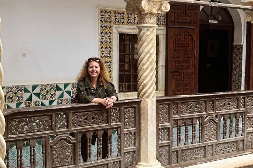 A person leaning on an ornate balcony railing with intricate designs and a patterned wall in the background. 1