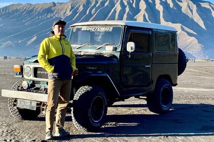 A person standing next to a black off-road vehicle with a license plate, in a desert landscape with mountains in the background. 2