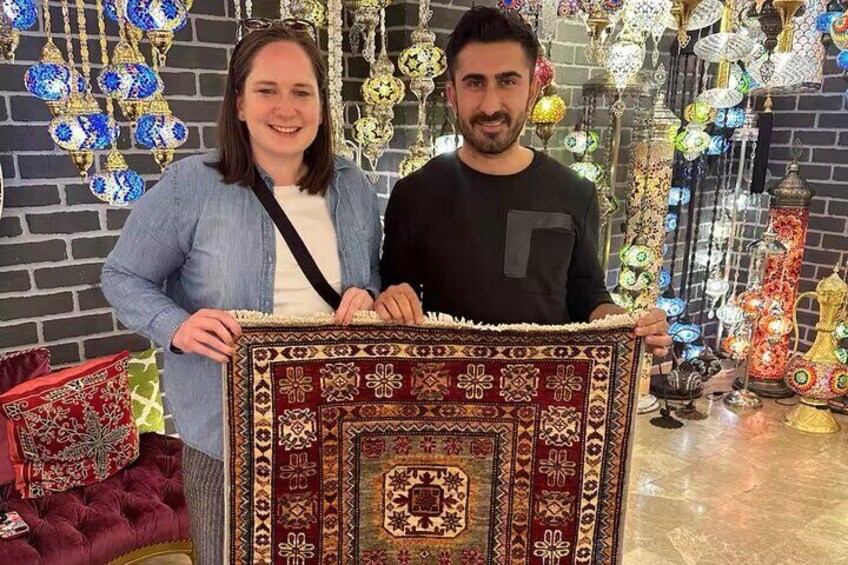 Two people holding a patterned rug in front of a wall decorated with hanging ornaments. 3