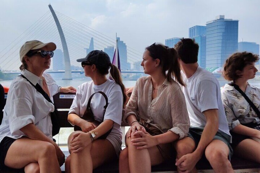 A group of people sitting on a boat with a bridge and buildings in the background. 2