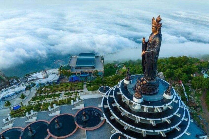 A statue on a circular platform with a surrounding garden, overlooking a valley with a building in the background. 1