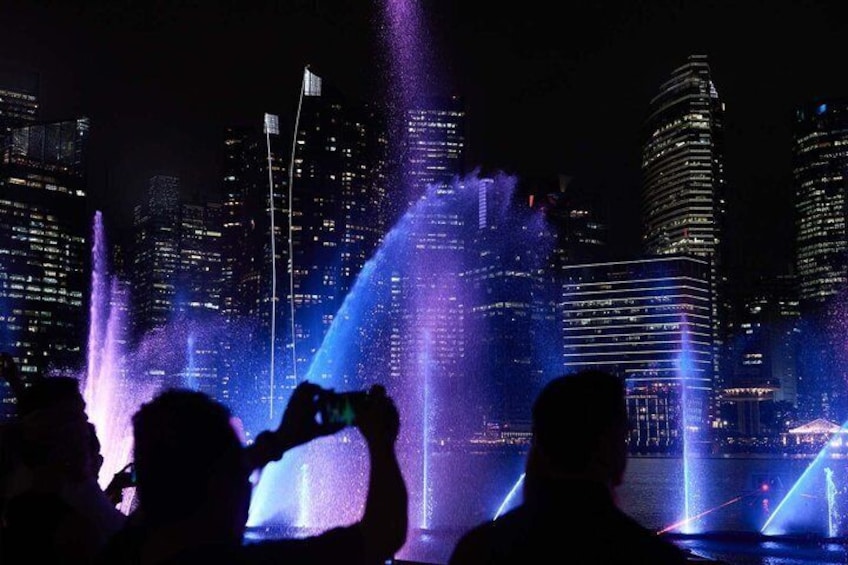 A nighttime cityscape with illuminated buildings and a water fountain display. 3