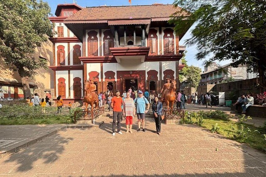 A historic building with a red-tiled roof and white walls, surrounded by a paved area with people walking and horse statues. 1
