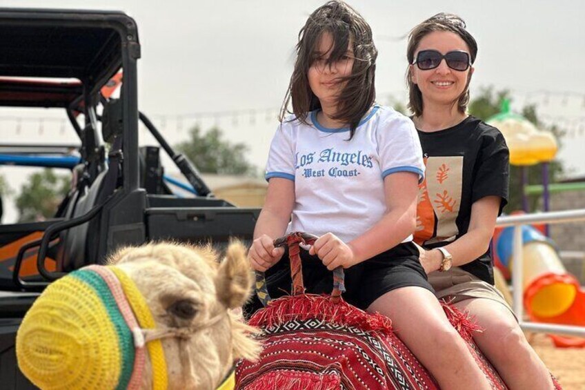 Two people riding a camel with a red saddle, a trailer, and a playground in the background. 2