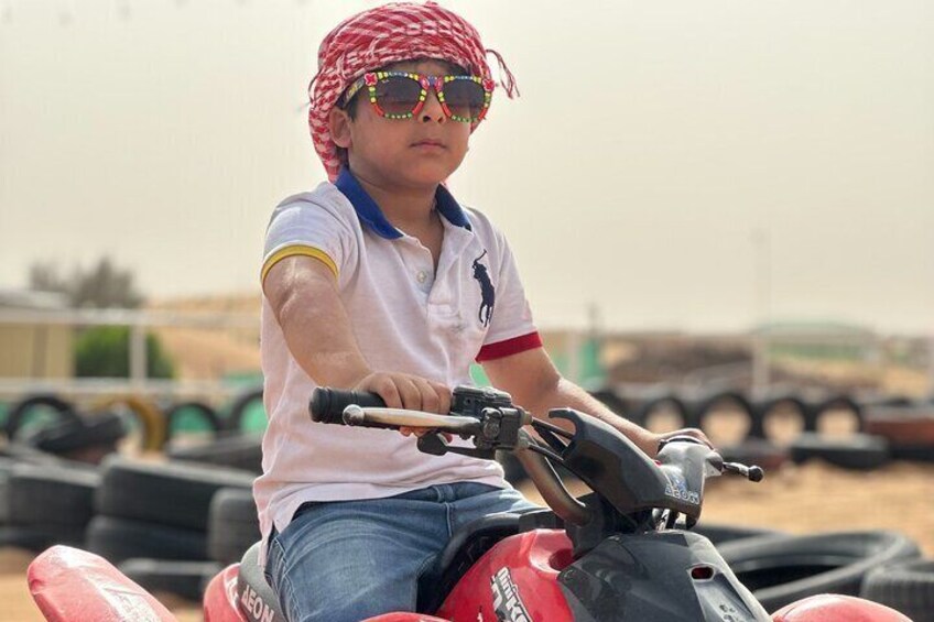 A child riding a red ATV in a desert-like setting with tire tracks and a vehicle in the background. 3