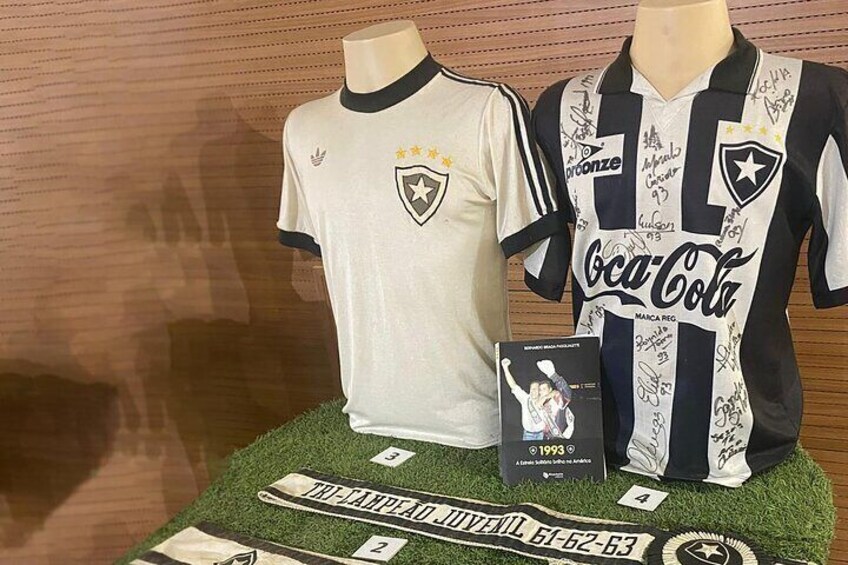 Two soccer jerseys on mannequins, one with a white and black design and the other with a white and blue design, both with various signatures. 3