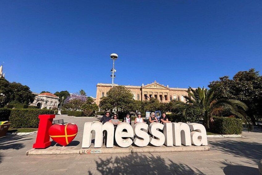 A large sign with the word 'messina' and a red cross symbol. In the background, there is a building with columns and a clear blue sky. 4