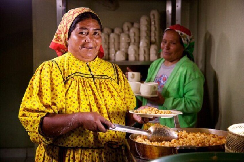 Two women serving food in a kitchen, one holding a ladle and the other a tray with bowls. 2