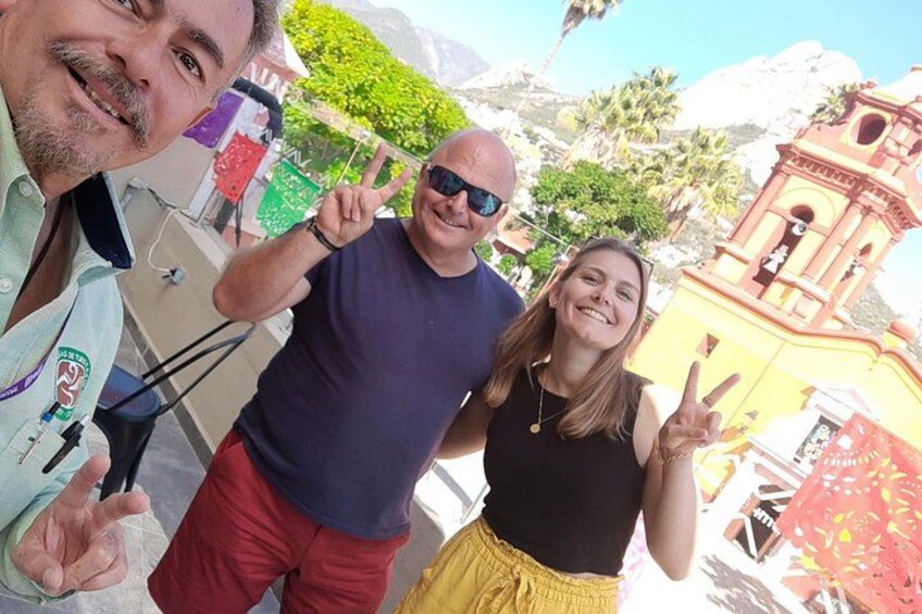 Three people posing with a camera, a bell tower, and greenery in the background. 3