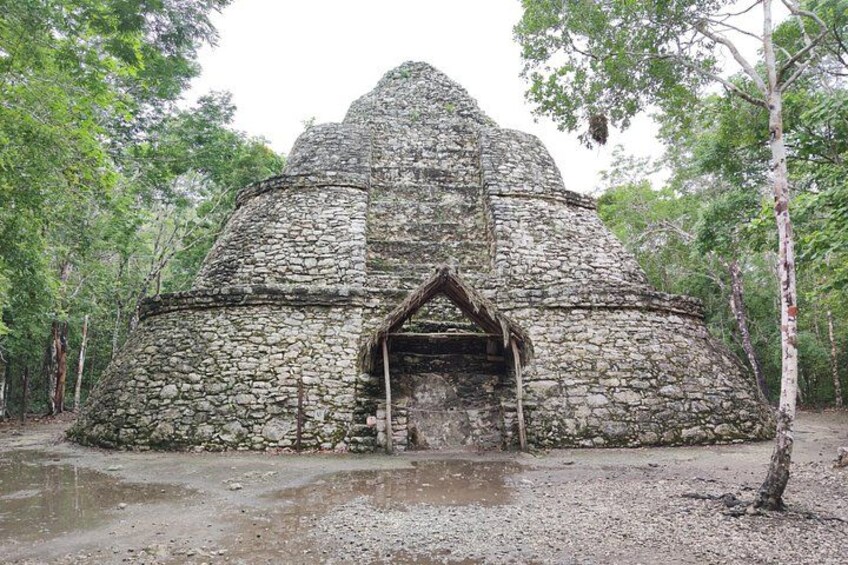A stone pyramid structure with a central entrance, surrounded by trees and a wet ground. 5