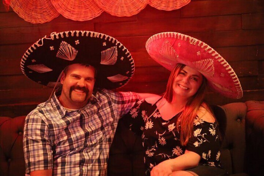 Two people wearing large sombreros, seated in a room with wooden walls and red lighting. 3