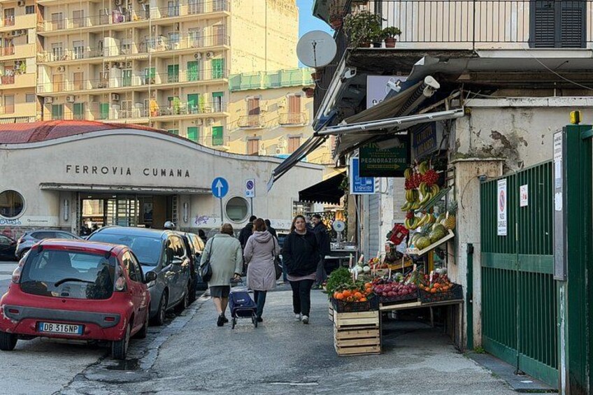 A street scene with parked cars, a market stall with fresh produce, and a building with a sign reading 'FERROVIA CUMANA'. 2