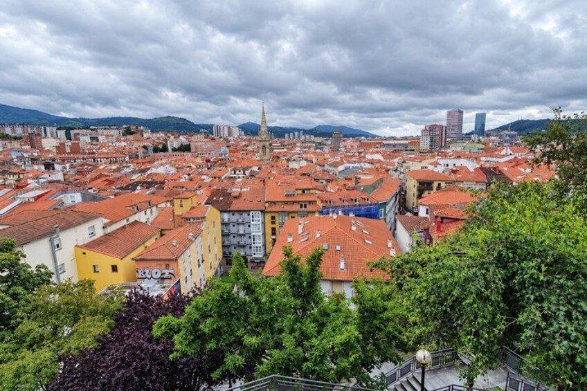 A cityscape with red-roofed buildings, a prominent tower, and green trees. 5