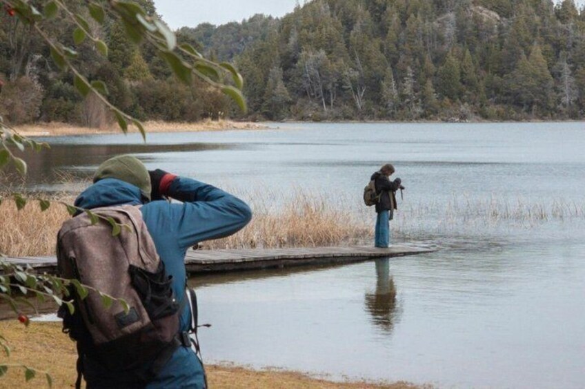 A person standing on a dock by a calm lake, wearing a backpack and a hat, with trees and a forest in the background. 2