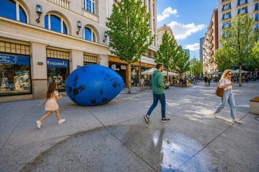 A public square with a large blue egg sculpture, storefronts, and pedestrians. 5