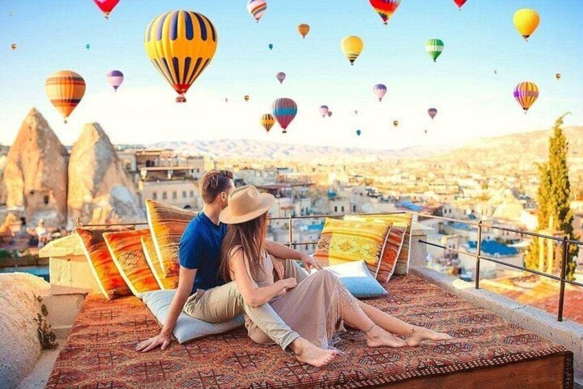 A couple sitting on a bed overlooking a cityscape with hot air balloons in the sky. 5