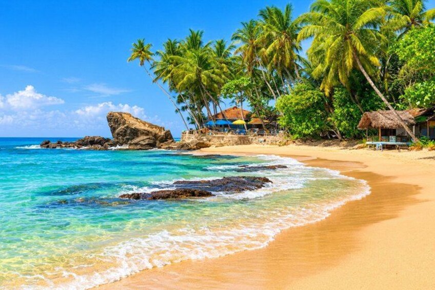A tropical beach with clear turquoise water, palm trees, and a rocky outcrop. 1