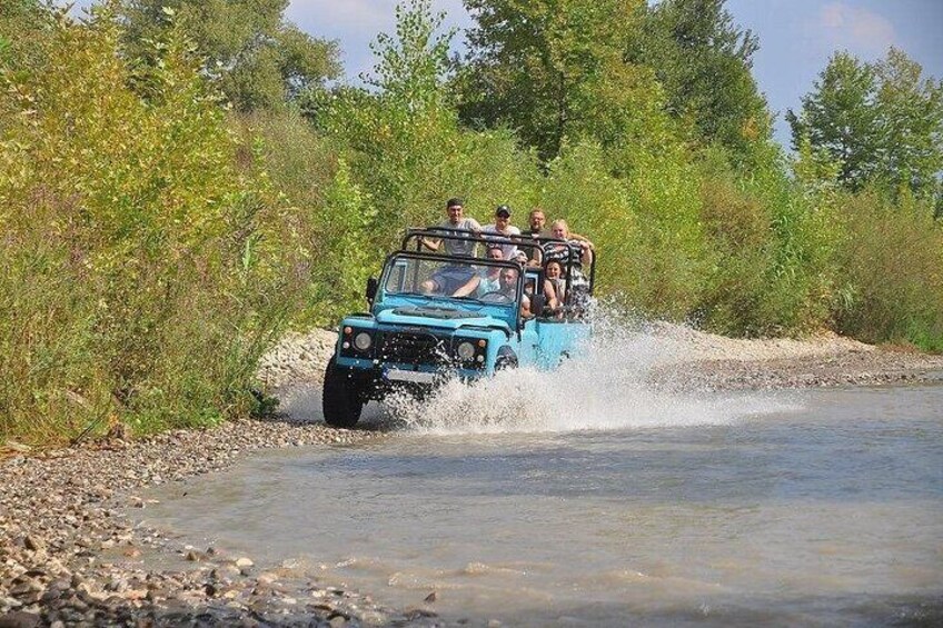 A blue off-road vehicle driving through a shallow water crossing surrounded by greenery. 4