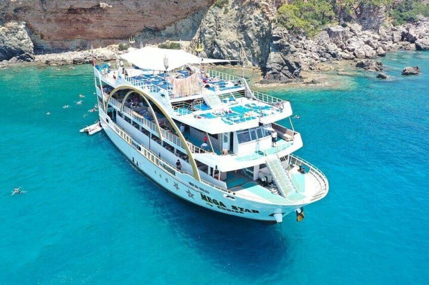 A large white boat with multiple decks and a blue canopy, floating in turquoise waters near rocky cliffs. 5