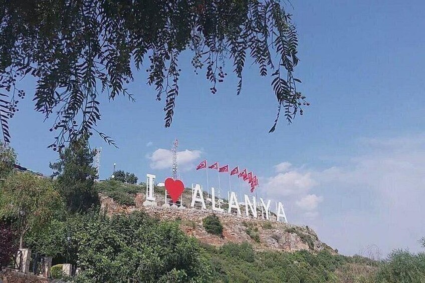A hilltop sign with the word 'ALANYA' and a red heart symbol, surrounded by greenery and flags. 4