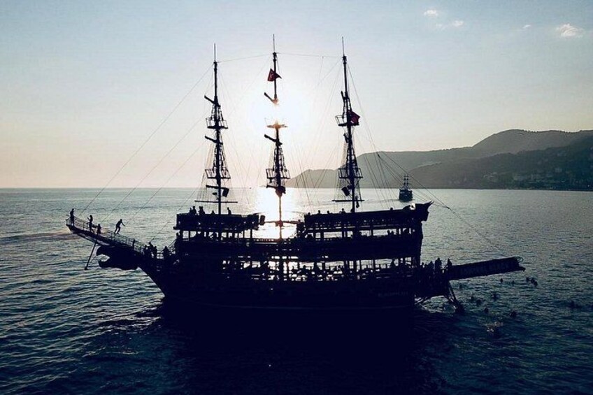 A large sailing ship with multiple masts and a crew on deck, anchored in calm waters with a hilly landscape in the background. 2