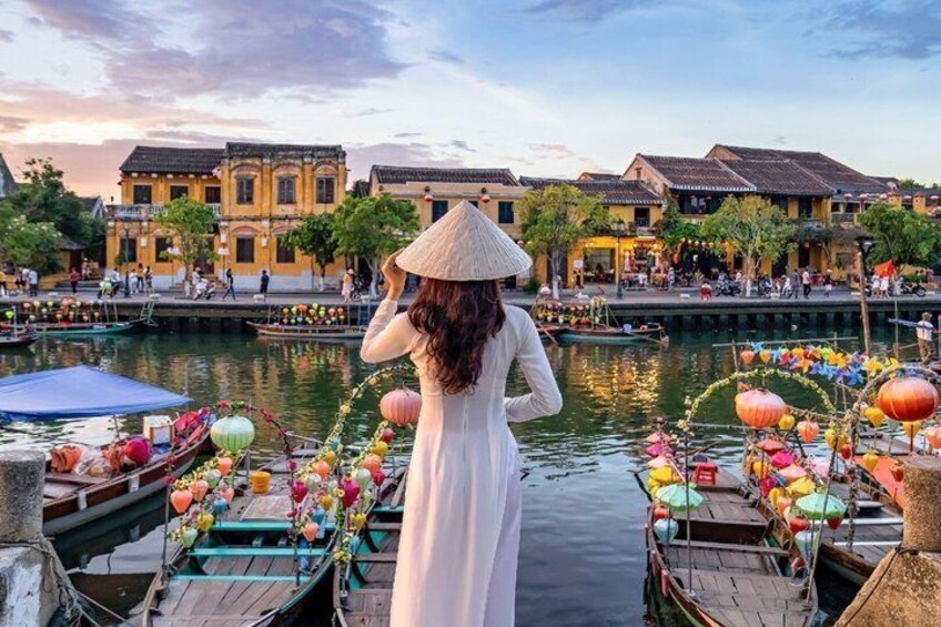 A person in a conical hat standing on a boat, a river with traditional buildings, and colorful lanterns. 1