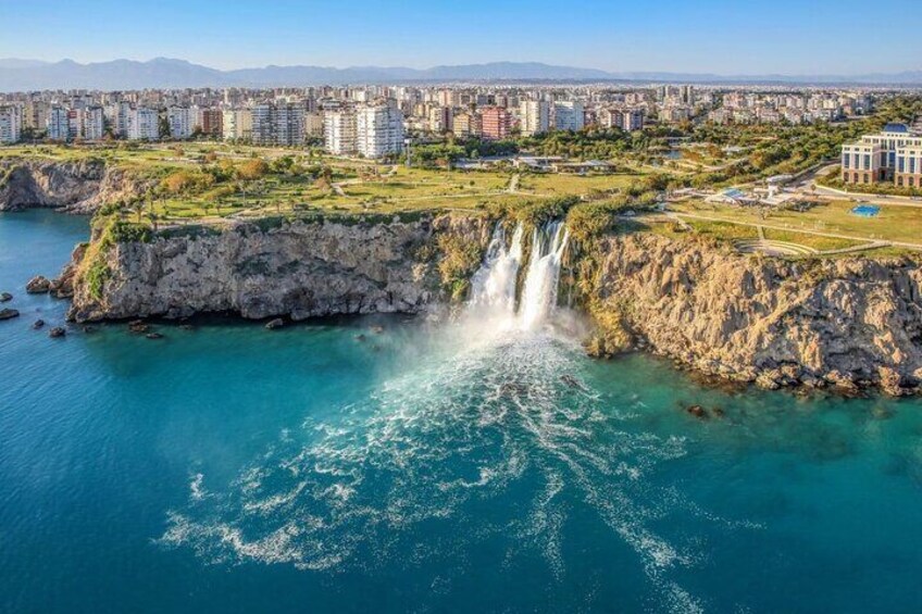 A coastal cityscape with a prominent waterfall, urban buildings, and a clear sky. 1