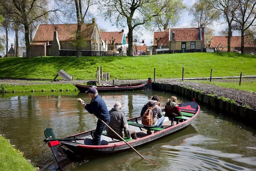 A narrow canal with a rowboat being rowed by a man, a small group of people in a rowboat, and a duck swimming in the water. 4