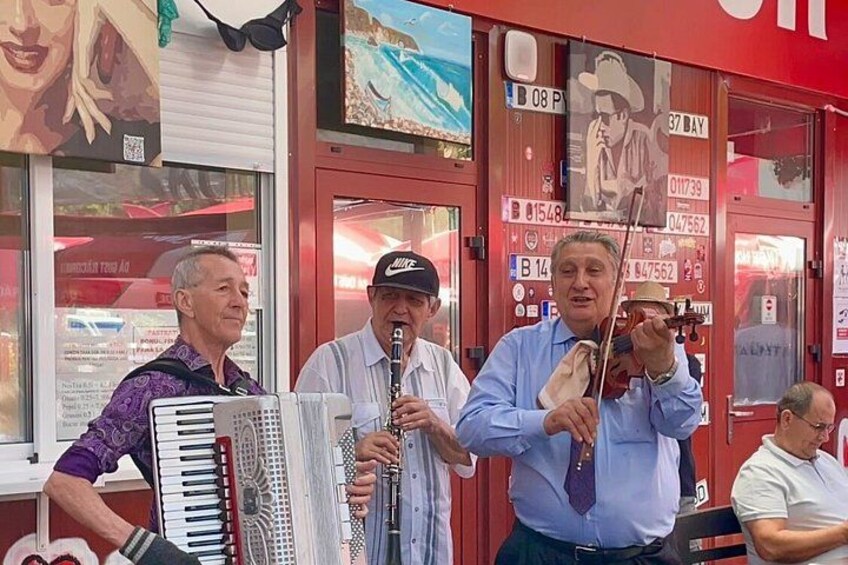 Three men playing musical instruments in front of a red building with various signs and artwork. 2