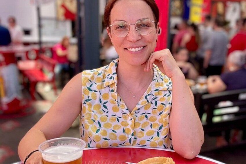 A person sitting at a table with a drink and food, in a busy outdoor setting with other diners. 1