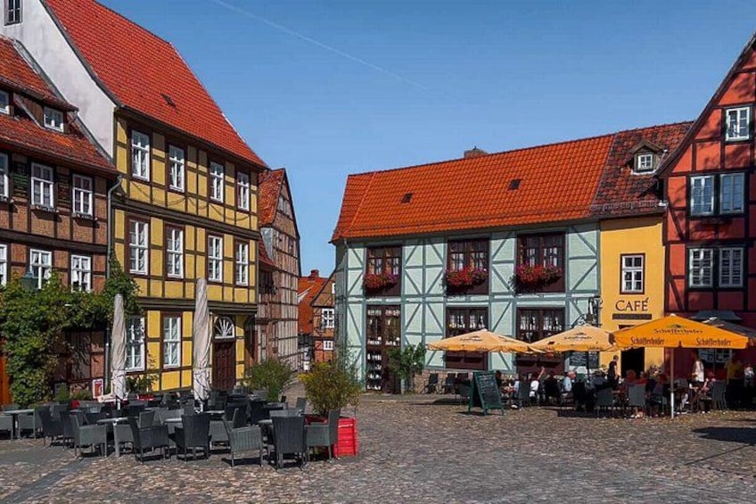 Una plaza adoquinada con edificios tradicionales de entramado de madera, mesas al aire libre en un café y un cielo azul despejado. 4