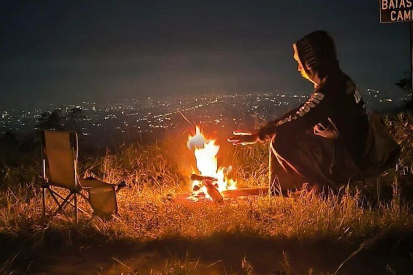 A person sitting by a campfire at night with a chair and a sign in the background. 5