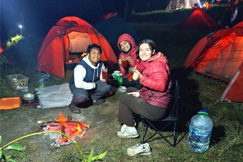 Three people sitting outdoors at night with red tents, a campfire, and a water bottle. 1