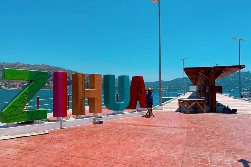 Large colorful letters spelling 'ZIHUA' on a waterfront promenade with a wooden shelter and a clear blue sky. 3